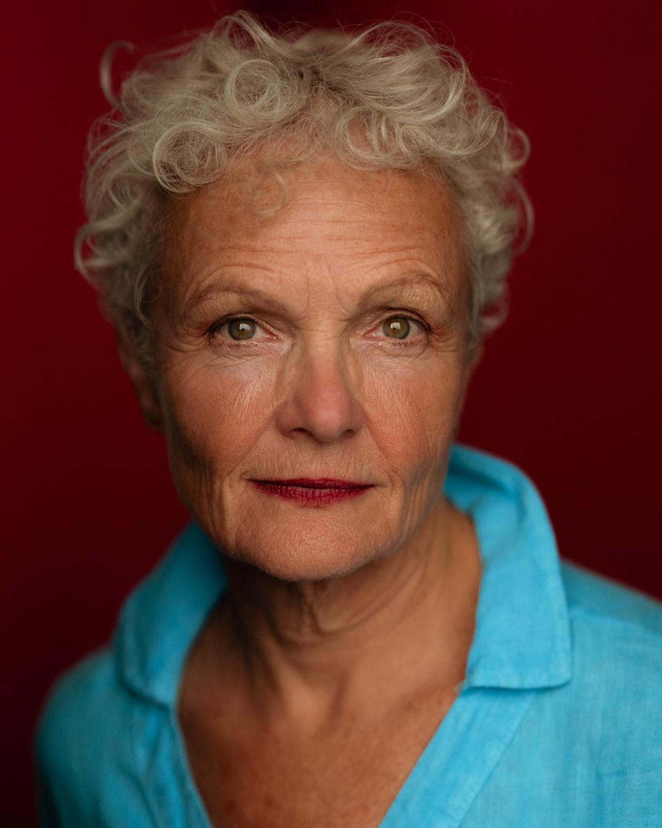 Elderly woman with short curly white hair and green eyes wearing a teal blouse against a red background.