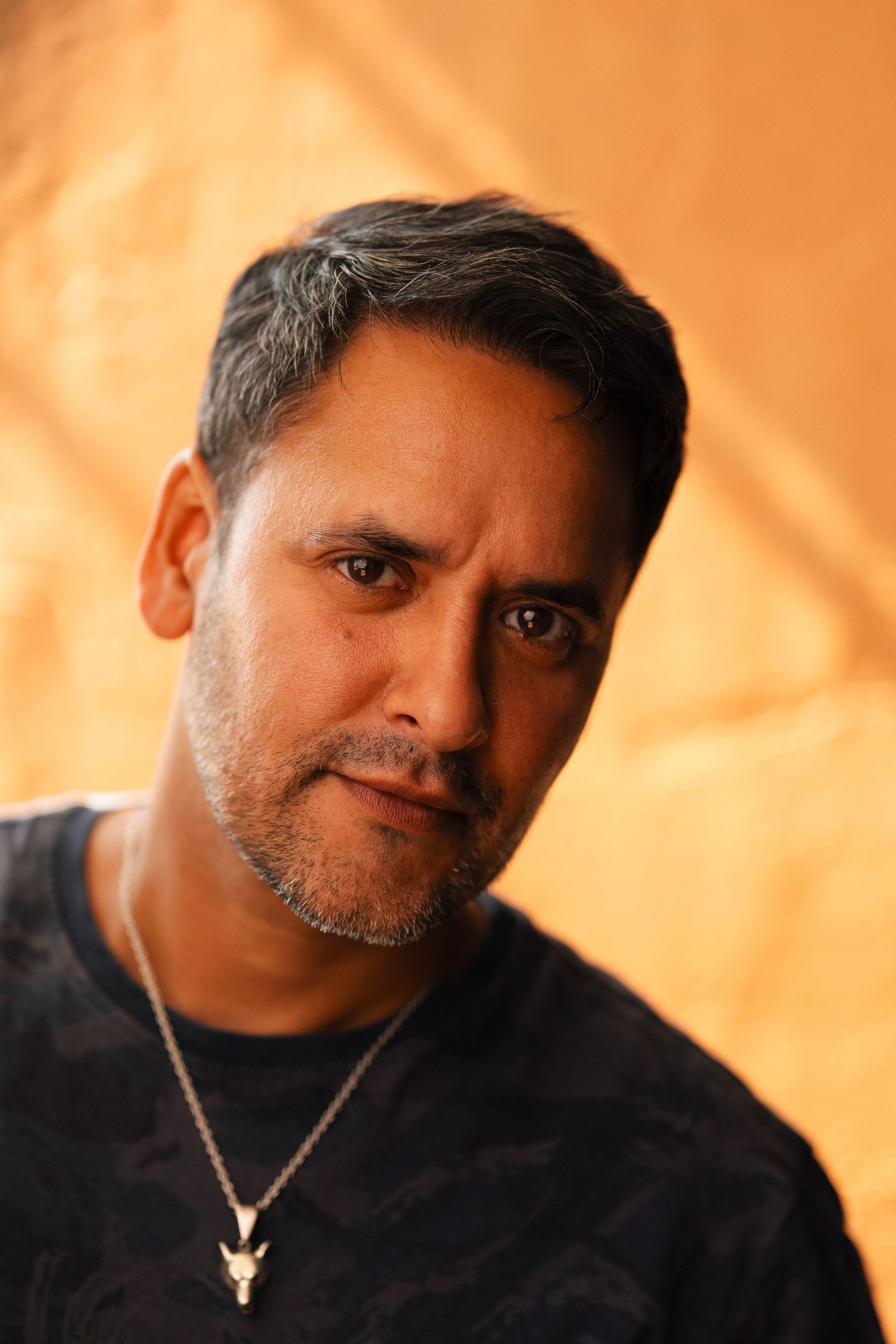Close-up portrait of a middle-aged man with dark hair, beard, and a bull skull necklace against an orange background.
