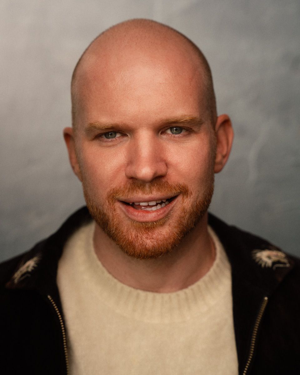 Bald man with red beard smiling, wearing cream sweater and dark jacket, professional headshot photo.