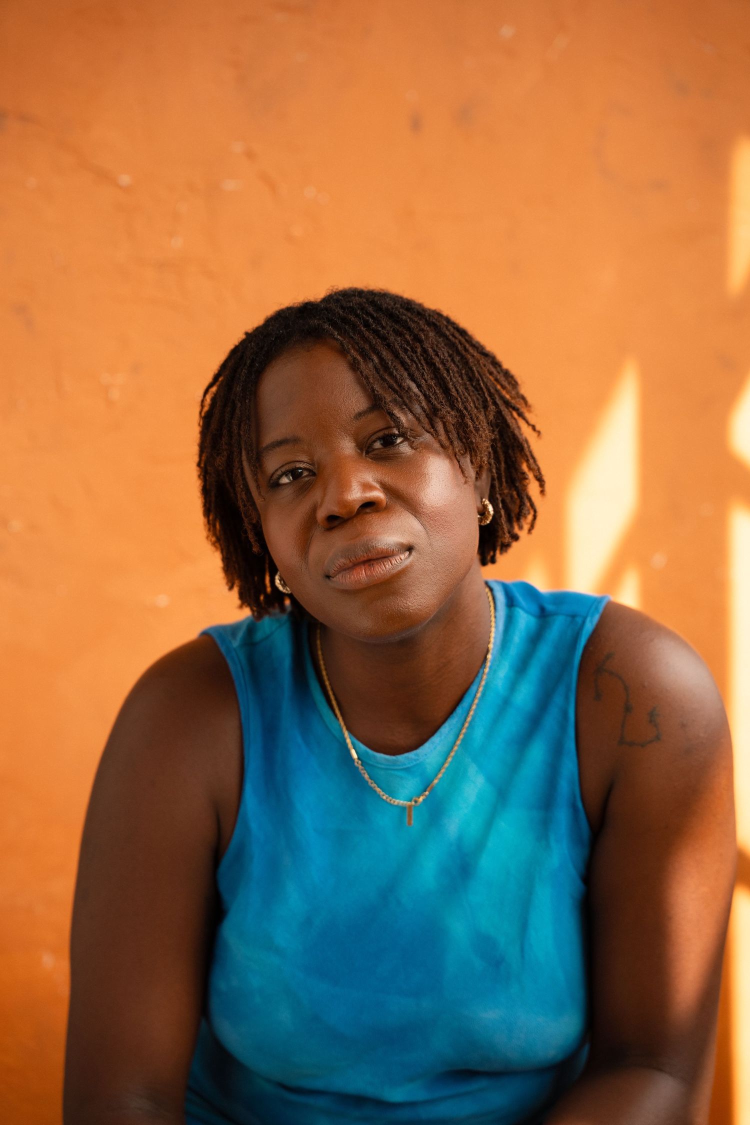 A woman with short dreadlocks wearing a blue sleeveless top and gold necklace poses against an orange wall.