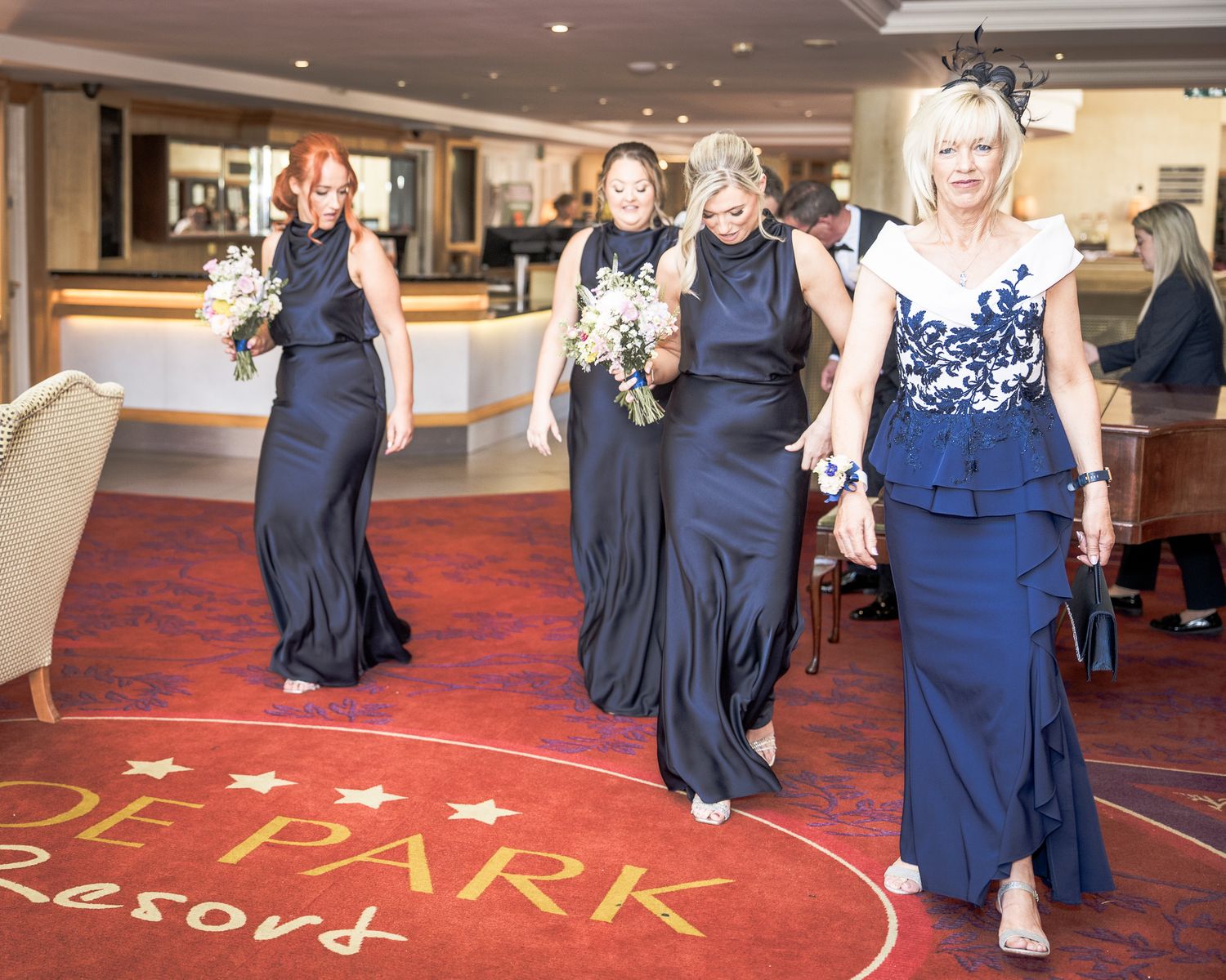 Bridesmaids in navy blue gowns walk through De Park Resort lobby carrying white bouquets at a wedding ceremony.
