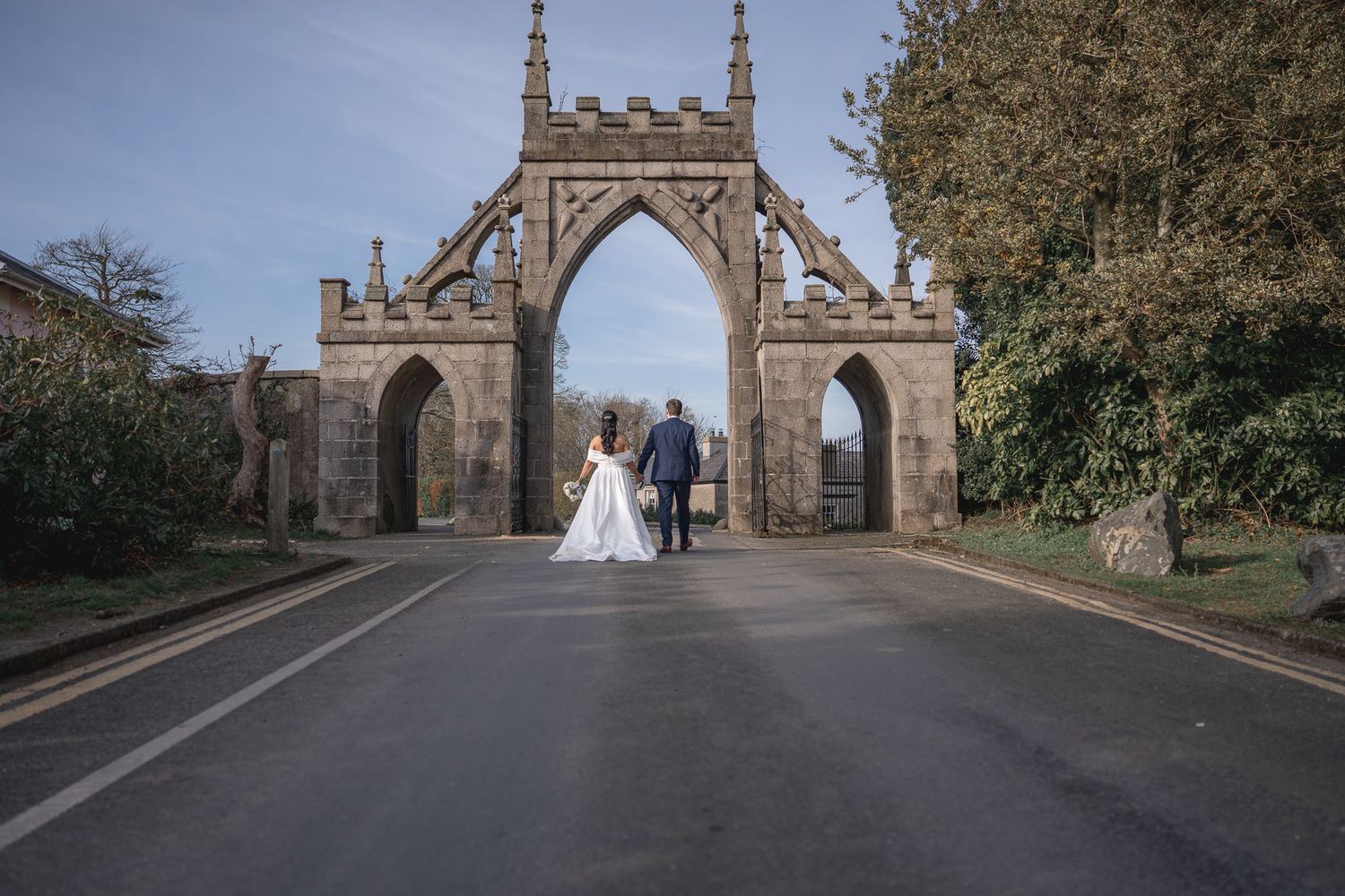 Bride and groom walking hand in hand through a grand Gothic stone arch gateway on a tree-lined road.