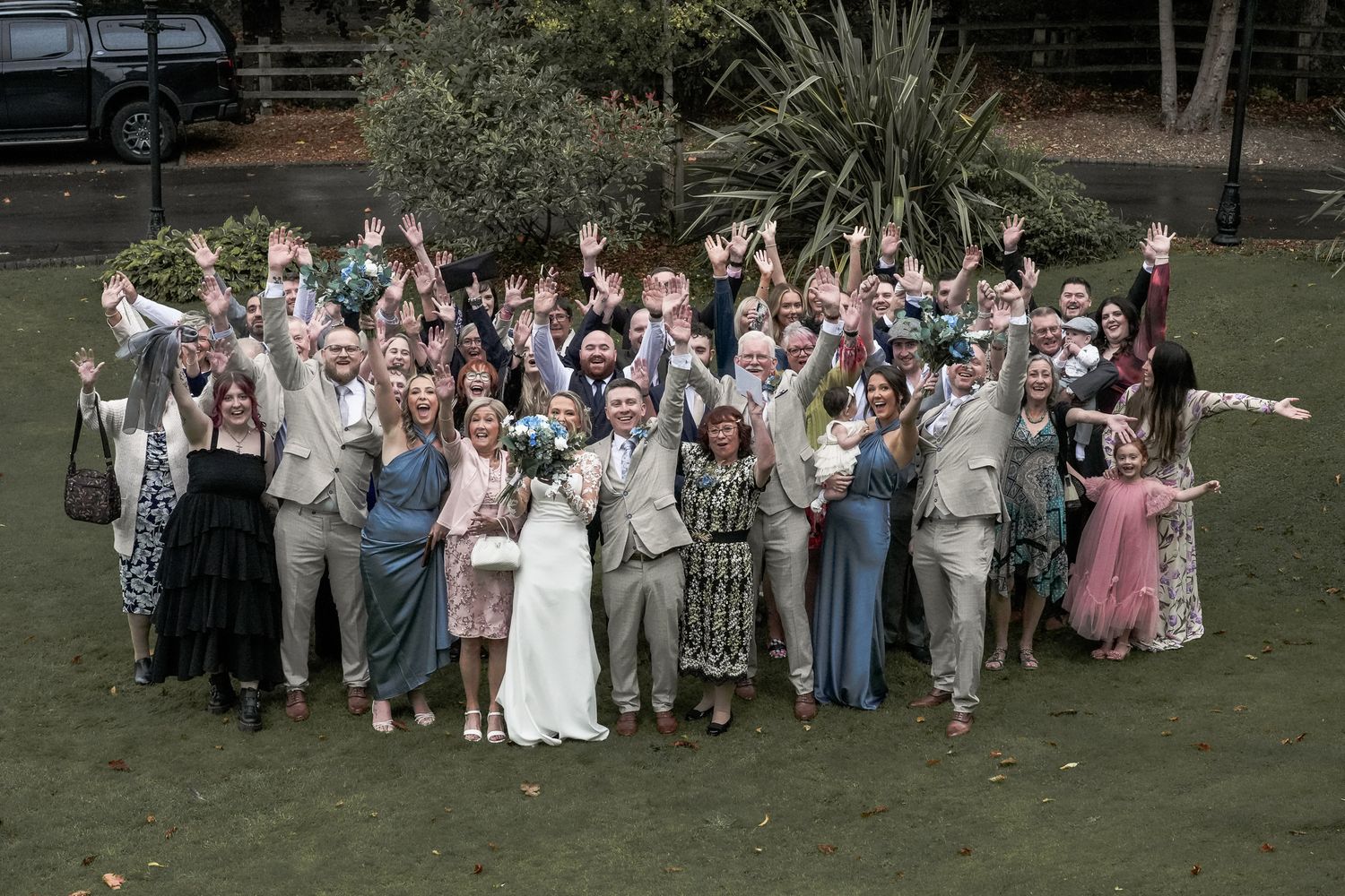 Large wedding group photo outdoors with guests cheering and raising hands, bride in white gown at center. Dave Martin wedding Photographer
