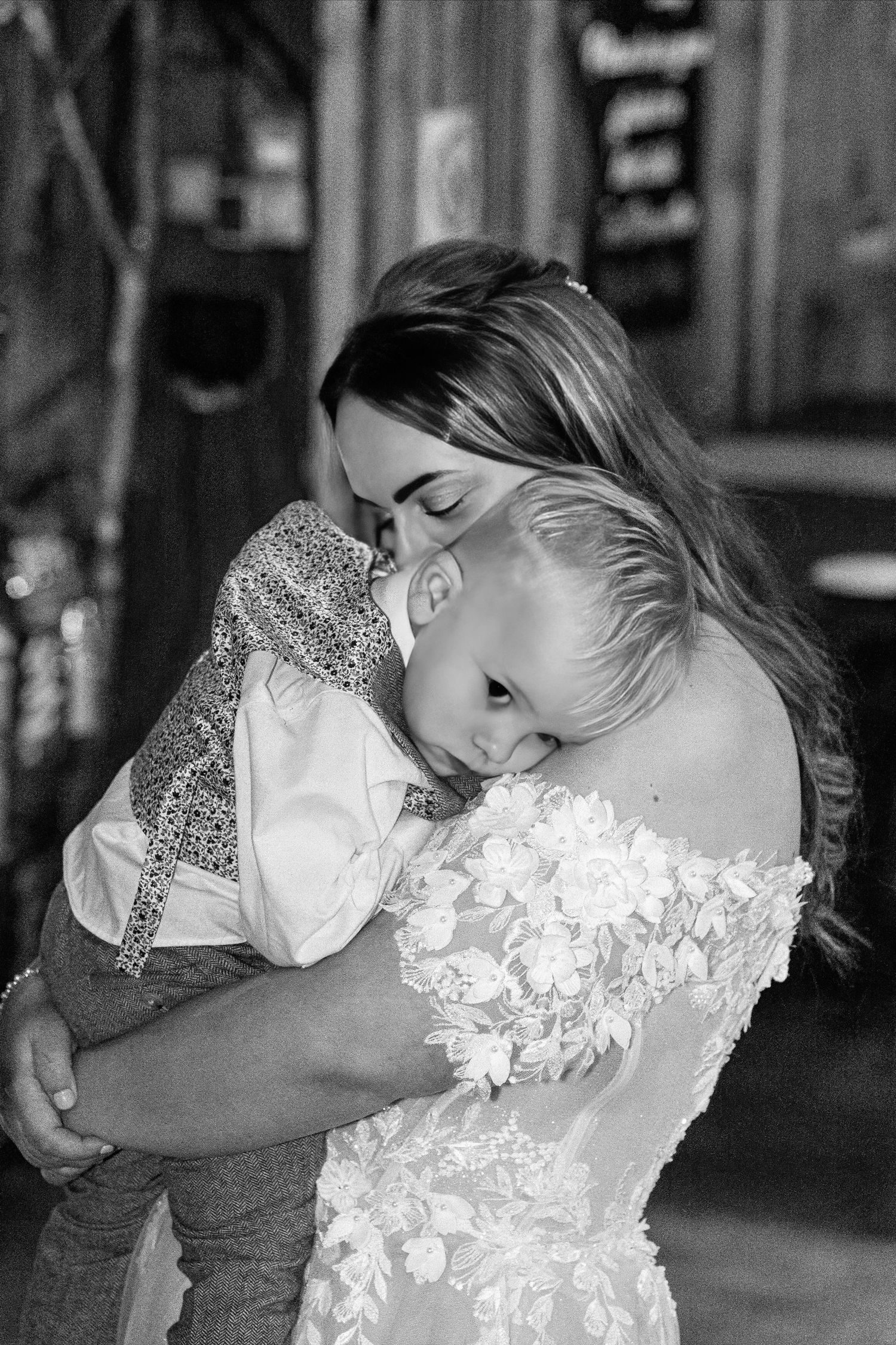 Bride in lace gown tenderly embraces a young child during an emotional wedding moment in black and white.