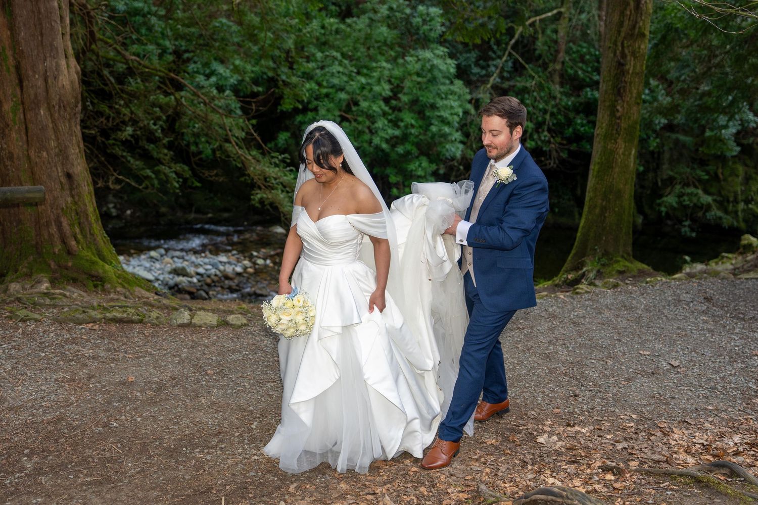 Groom in navy suit lifts bride's white wedding gown train as they walk along a forest path near a stream.