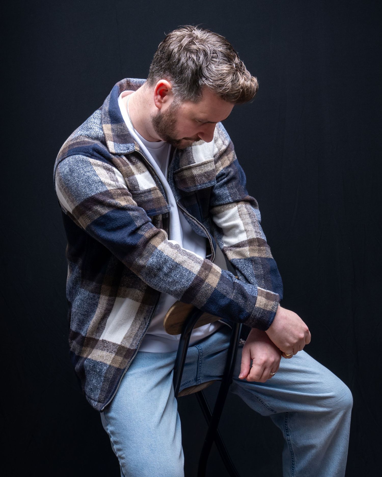 A bearded man in a plaid flannel shirt sitting on a stool against a dark background. Dave Martin Photography Studio Belfast