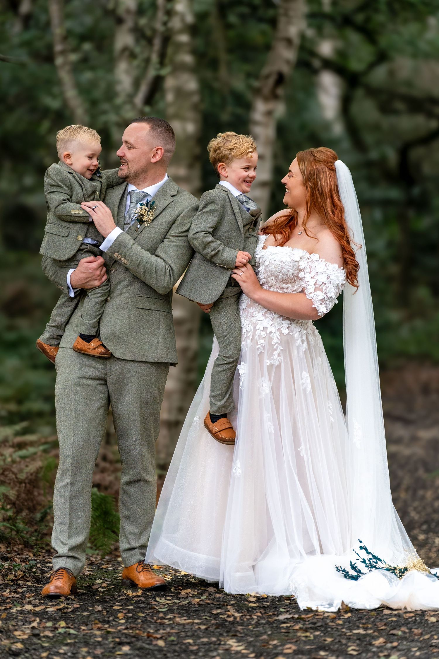 Bride and groom holding two young boys in matching grey suits, smiling together in a woodland setting. By Dave Martin Photography