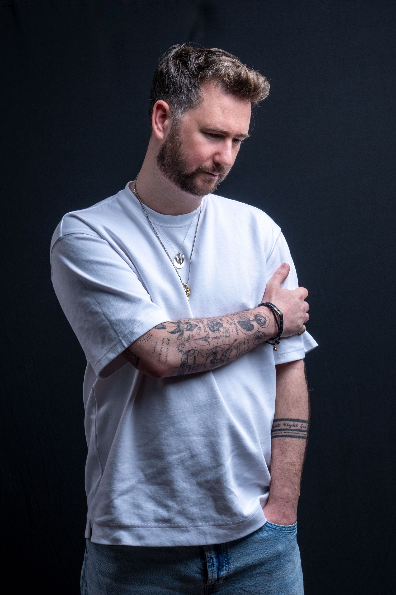 Tattooed man in white t-shirt and jeans poses against dark background, looking down with arms crossed. Dave Martin Photography Studio