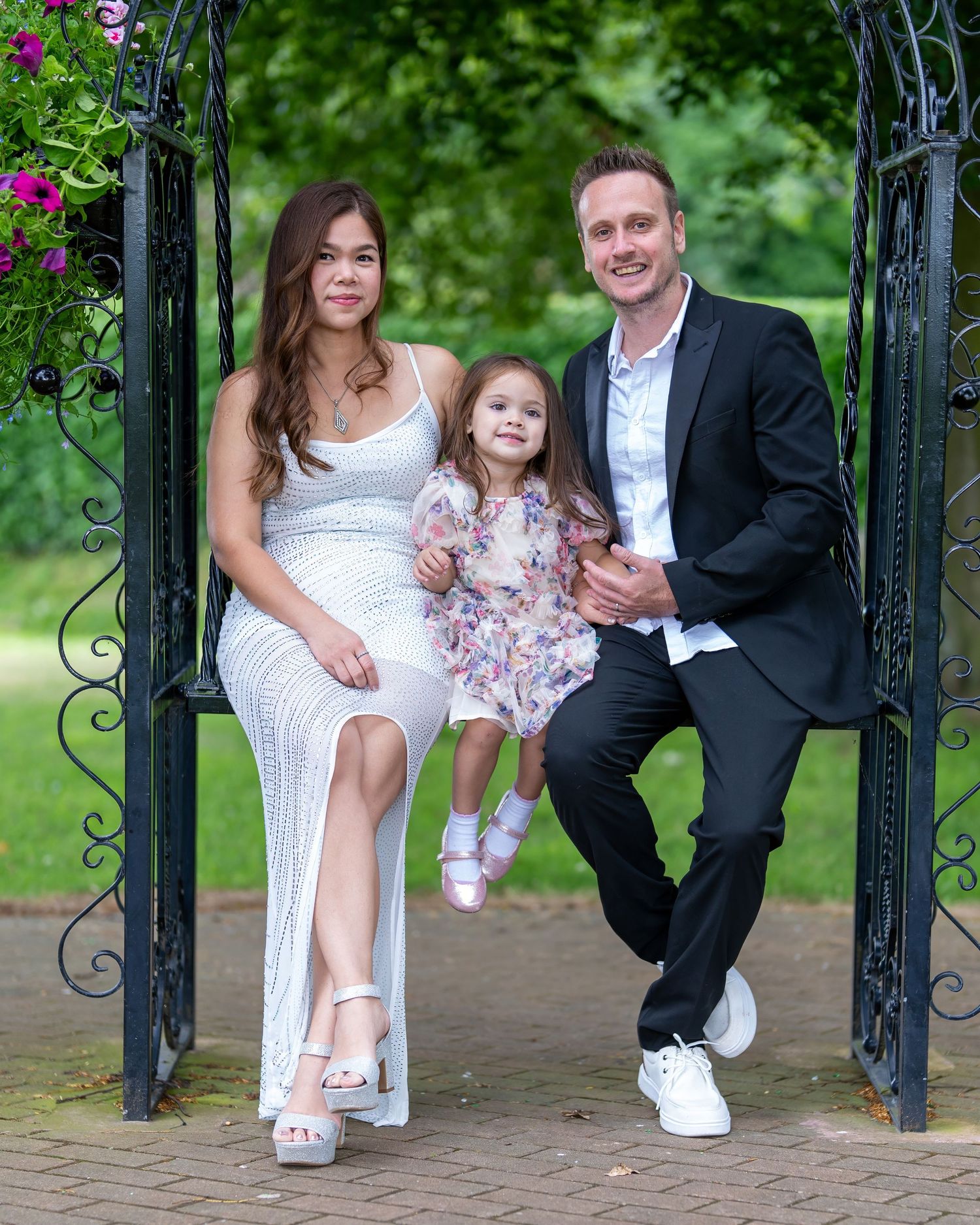 A smiling family of three poses under a garden archway, with a woman in white, a little girl, and a man in a suit. Dave martin wedding photo