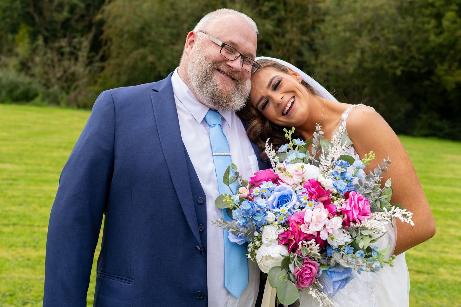 Bride smiling and leaning on older gentleman in blue suit, both holding colorful bouquet outdoors. Dave Martin Photographer Northern Ireland