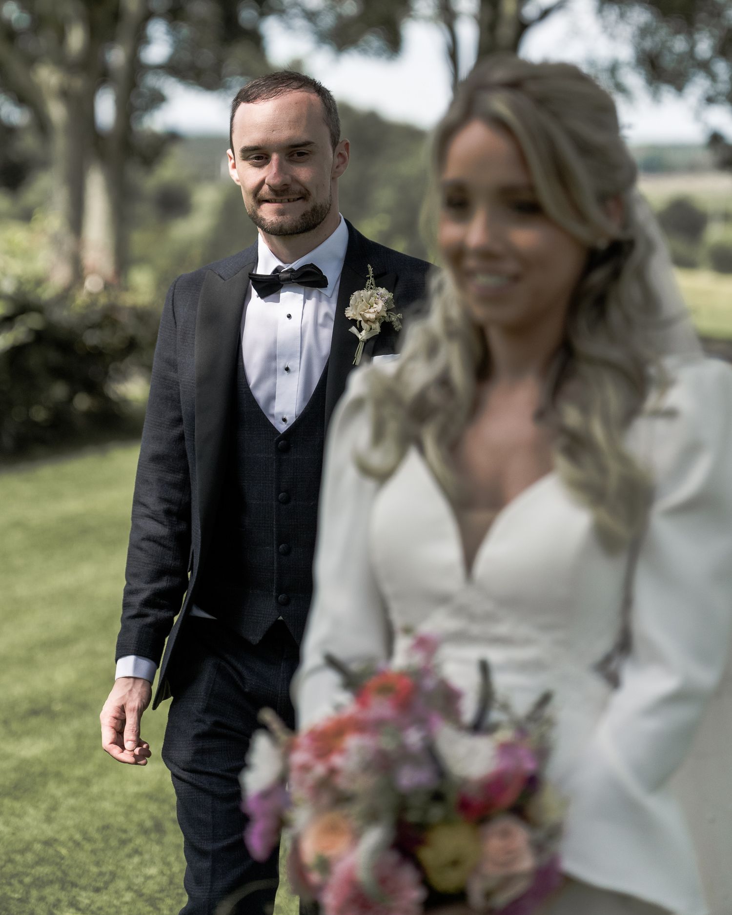 Groom in black tuxedo walks toward bride in white wedding dress holding colorful bouquet outdoors. Northern ireland weddings