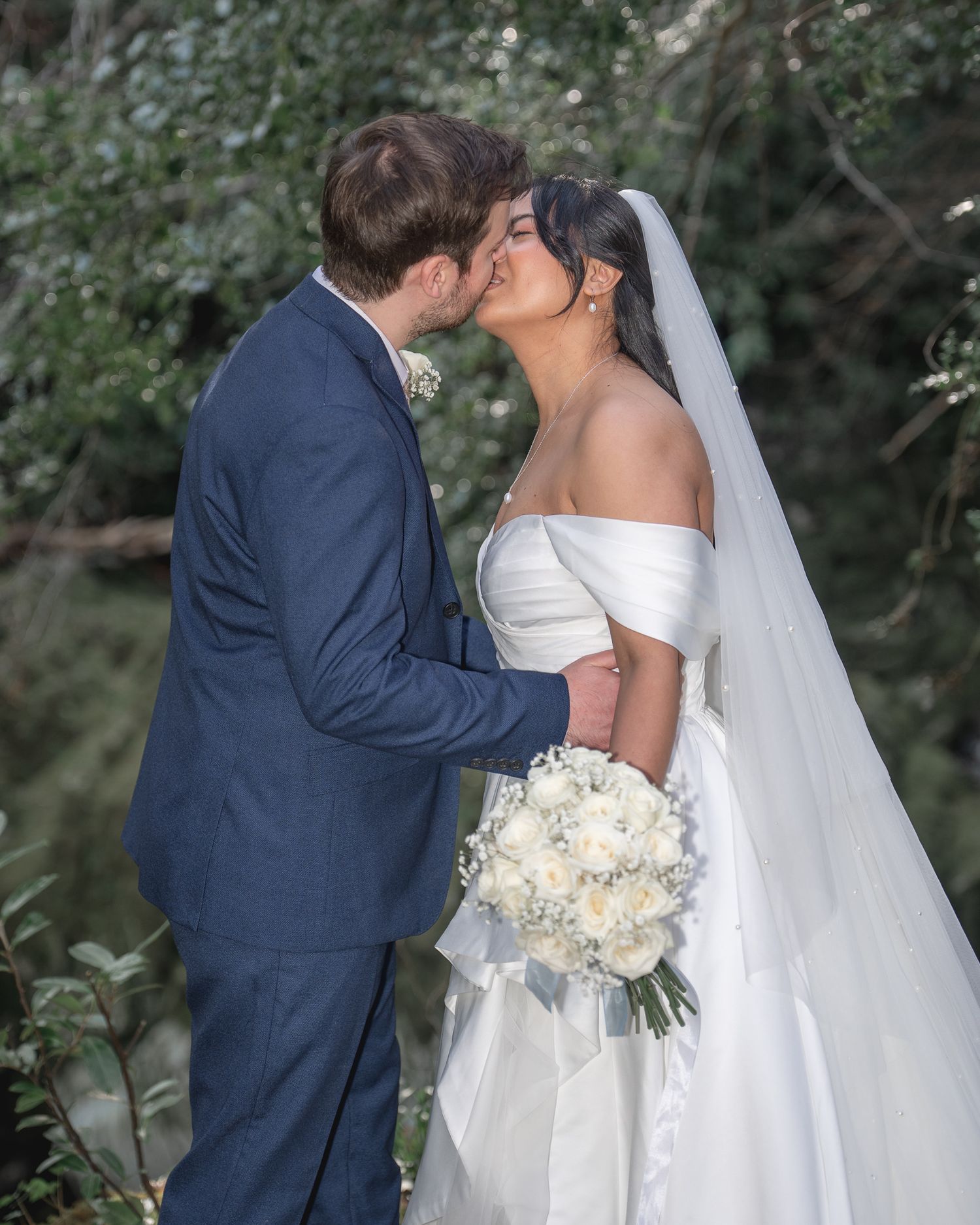A bride and groom share a romantic kiss outdoors, she holds a white bouquet wearing a strapless gown with veil.
