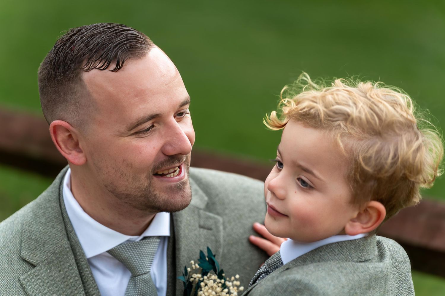 A smiling groom in a grey suit with boutonniere holds a curly-haired toddler boy at a wedding celebration.
