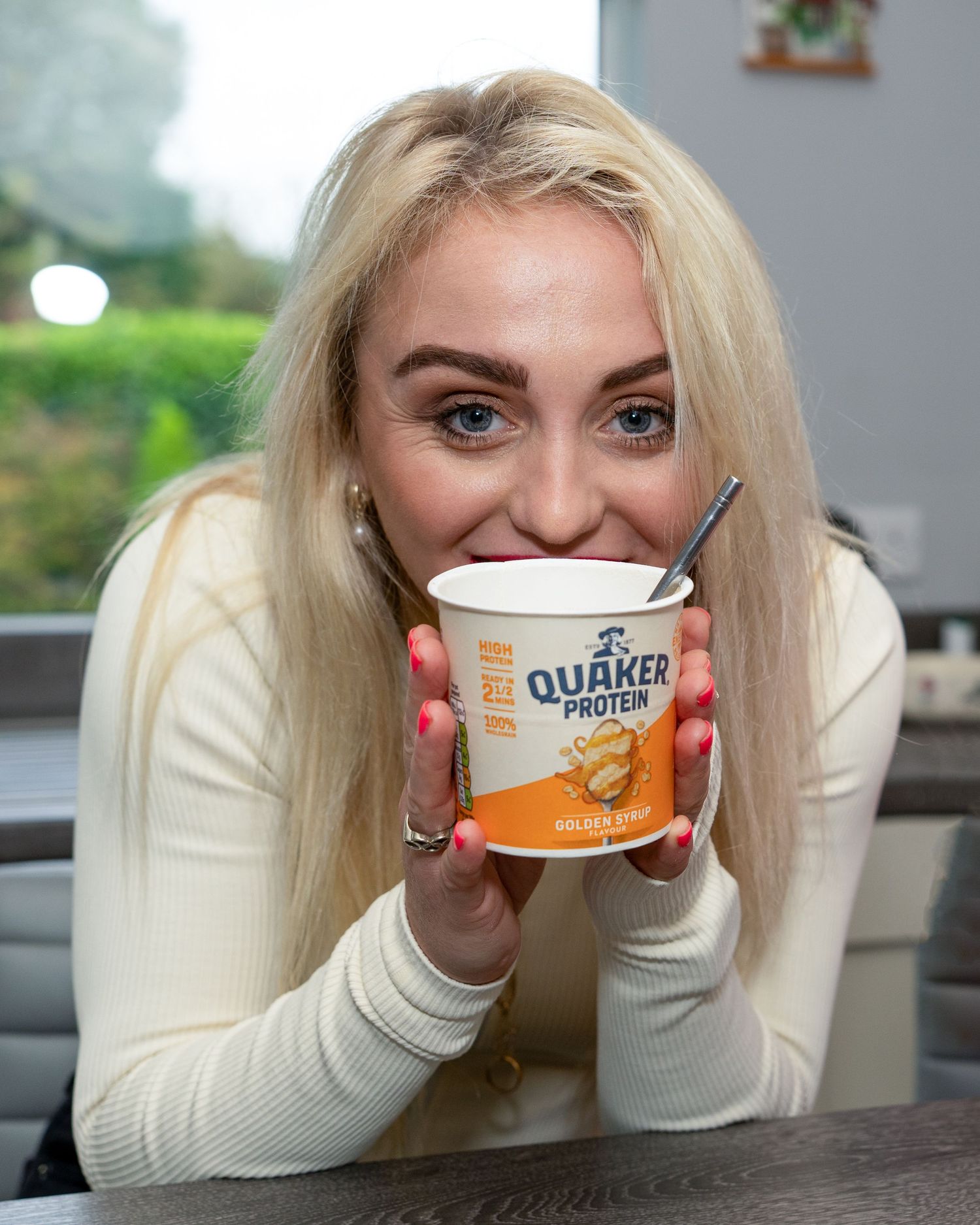 Smiling blonde woman holding a Quaker Protein Golden Syrup oat pot close to her face. Dave Martin Photography Studio Belfast Dave Martin cel