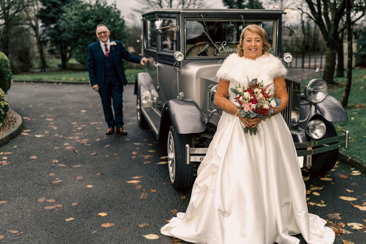 Bride holding bouquet beside vintage car with groom approaching on a tree-lined driveway. By Dave Martin Photography