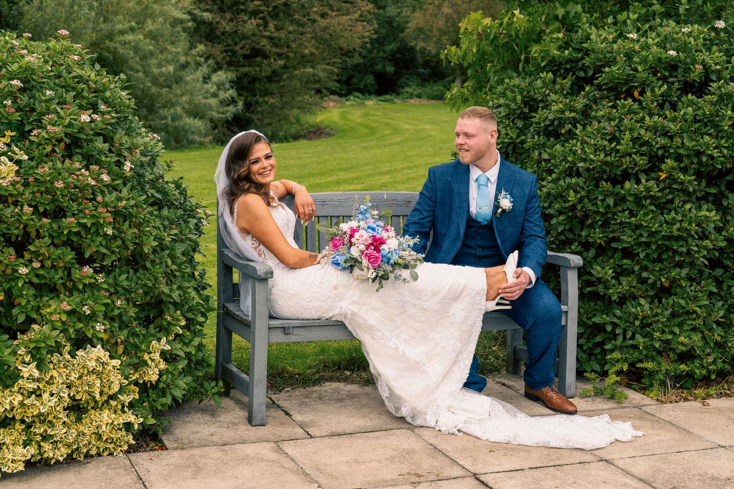 Bride and groom sitting on a garden bench, bride in lace gown holding colorful bouquet, groom in navy suit. By Dave Martin Photography