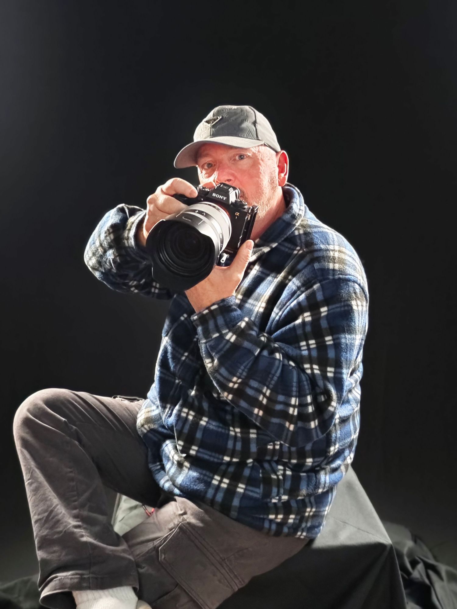 Man in plaid shirt and cap sits on ground aiming a professional DSLR camera with telephoto lens against dark background.
