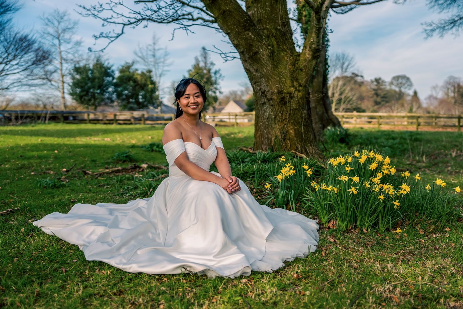 Bride in white off-shoulder gown sitting on grass near daffodils under a tree on a sunny spring day. Tollymore forest