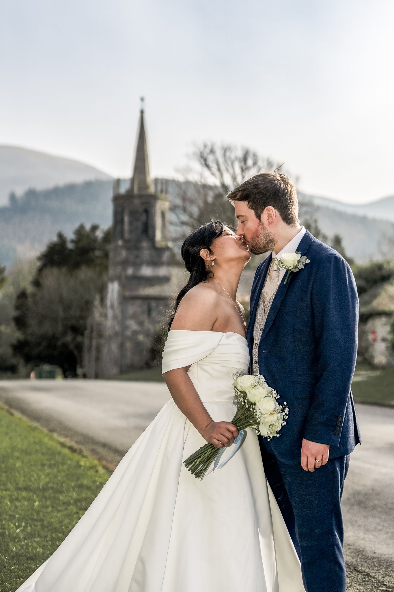Bride and groom share a romantic kiss outdoors, with a church steeple and mountains in the background on their wedding day.