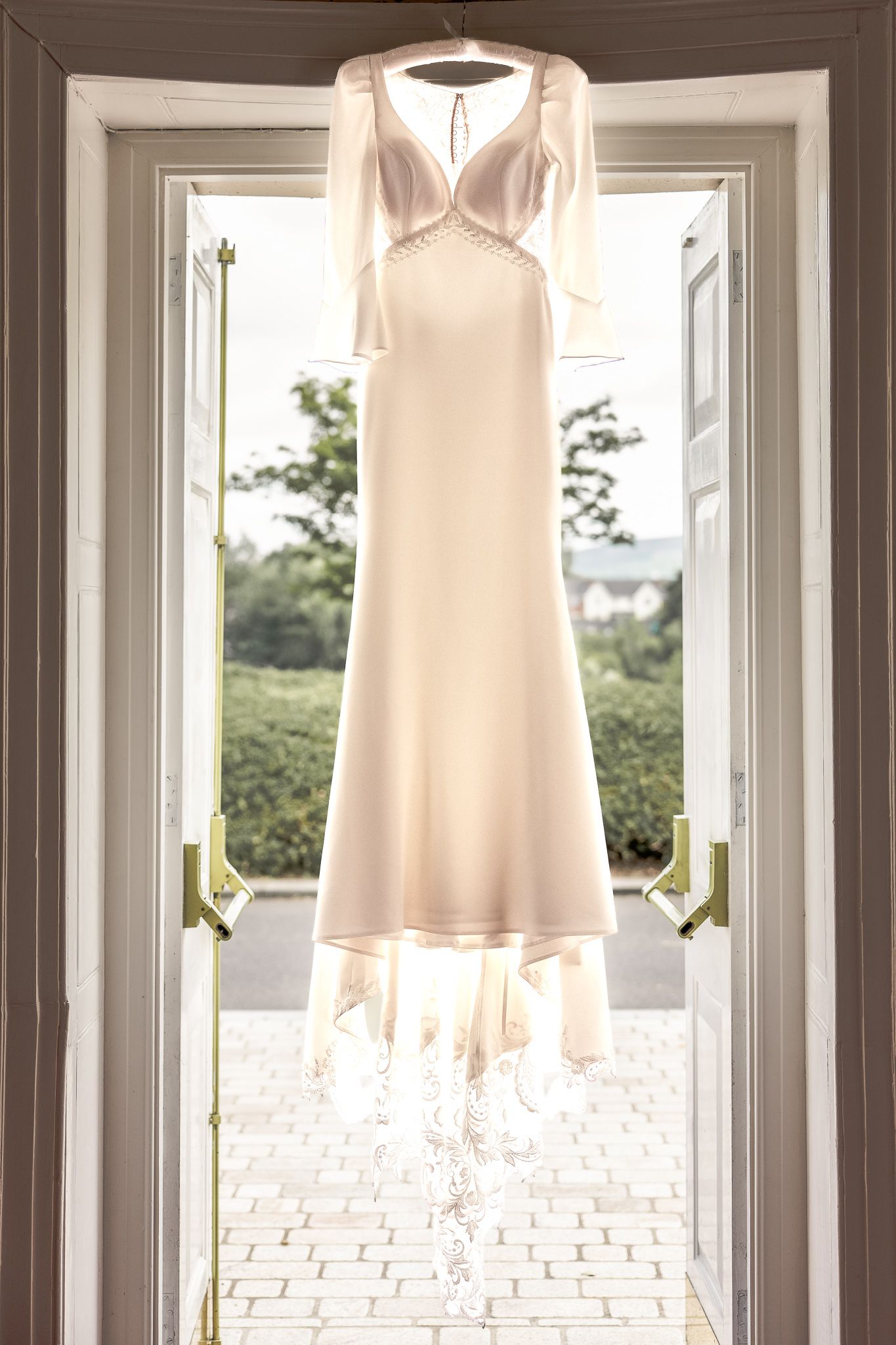 Elegant ivory wedding dress with lace hem hanging in open French doors with garden view. Dave Martin wedding Photographer NI