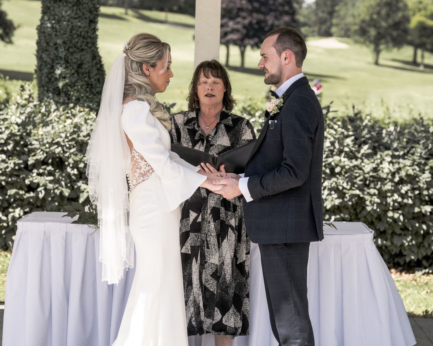 Bride and groom exchange vows with officiant outdoors at elegant wedding ceremony surrounded by greenery. Dave Martin wedding Photographer