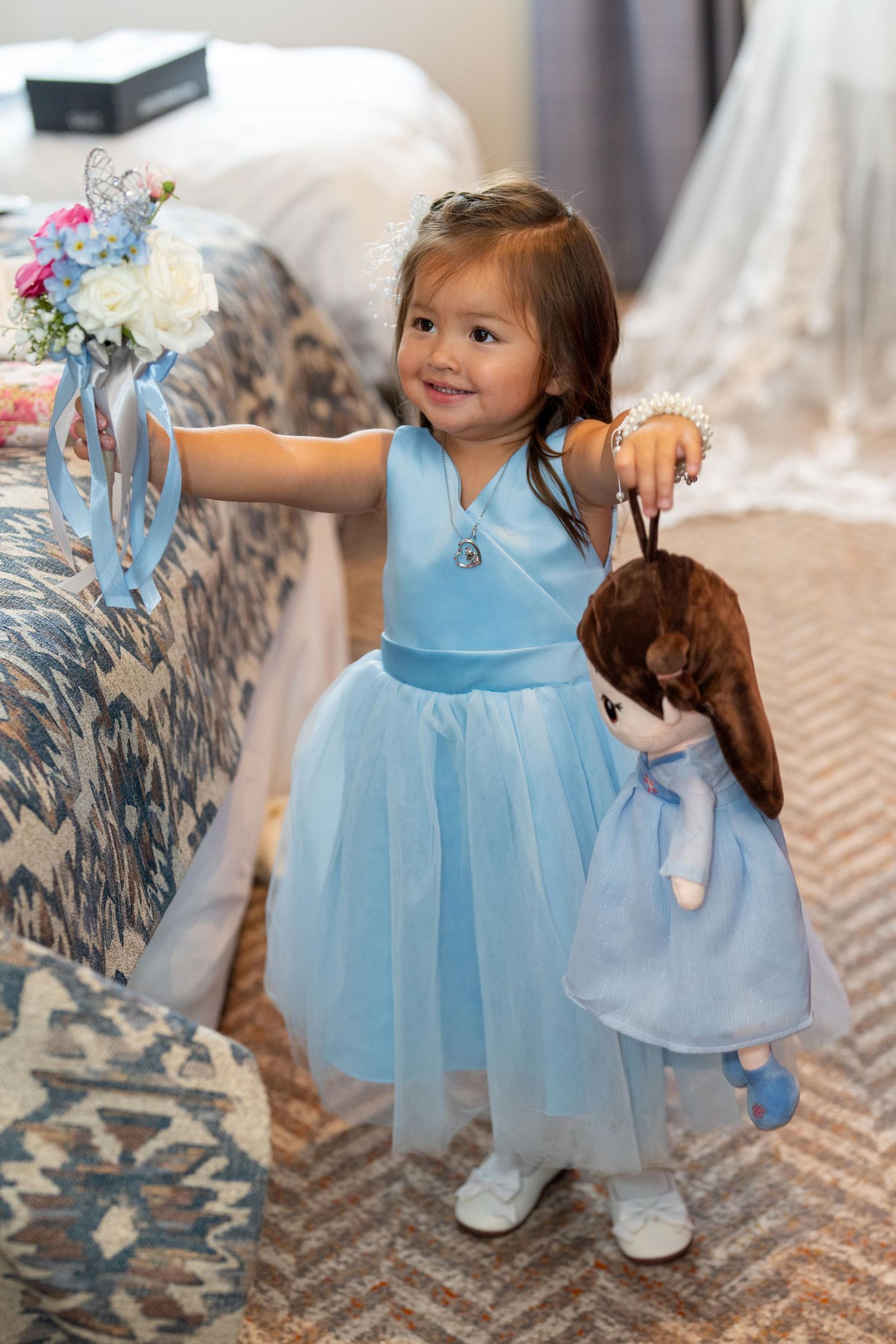 Smiling toddler girl in blue tulle dress holding stuffed animal and flowers at a wedding celebration.