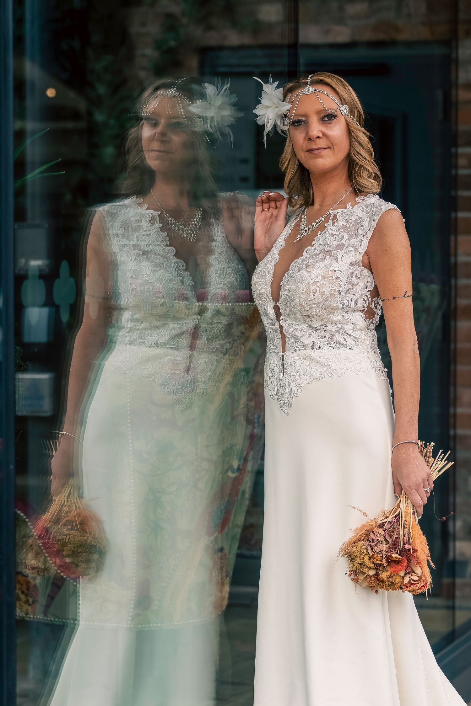 Elegant bride in lace wedding dress holding dried bouquet, reflected in glass door, wearing floral headpiece. By Dave Martin Photography