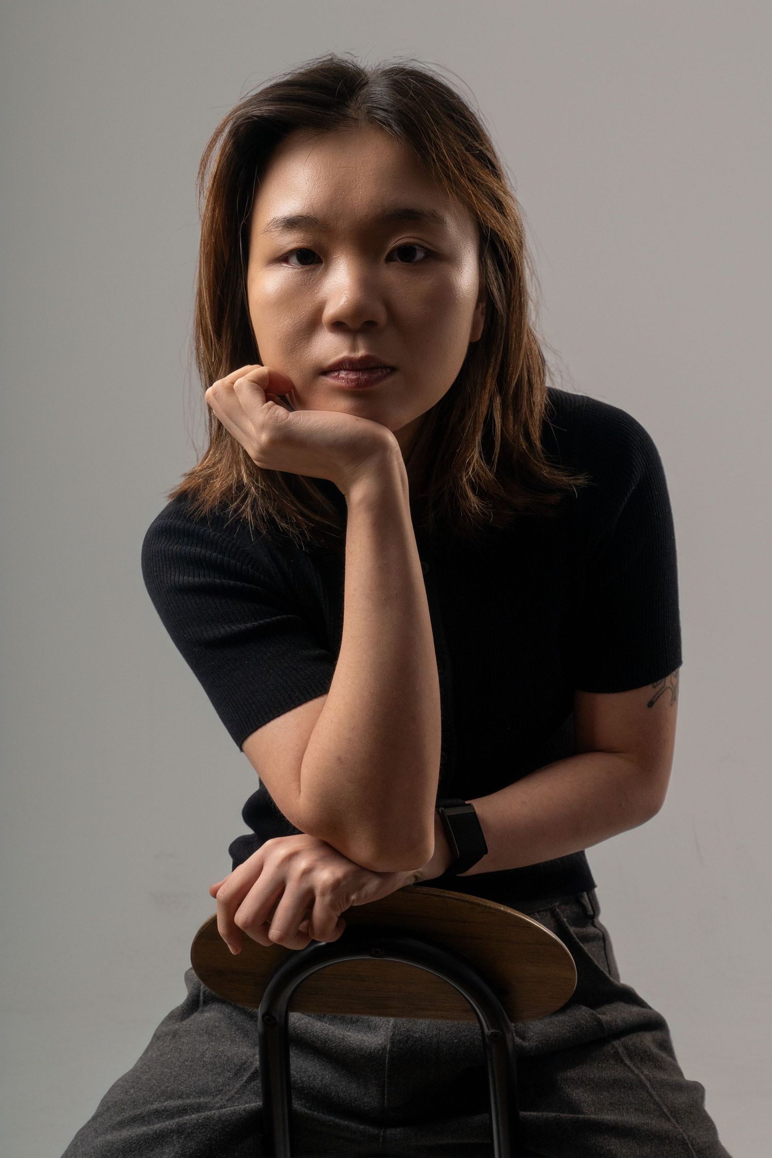 A young Asian woman in a black t-shirt sits confidently, resting her chin on her hand against a gray background. Dave Martin Photography.