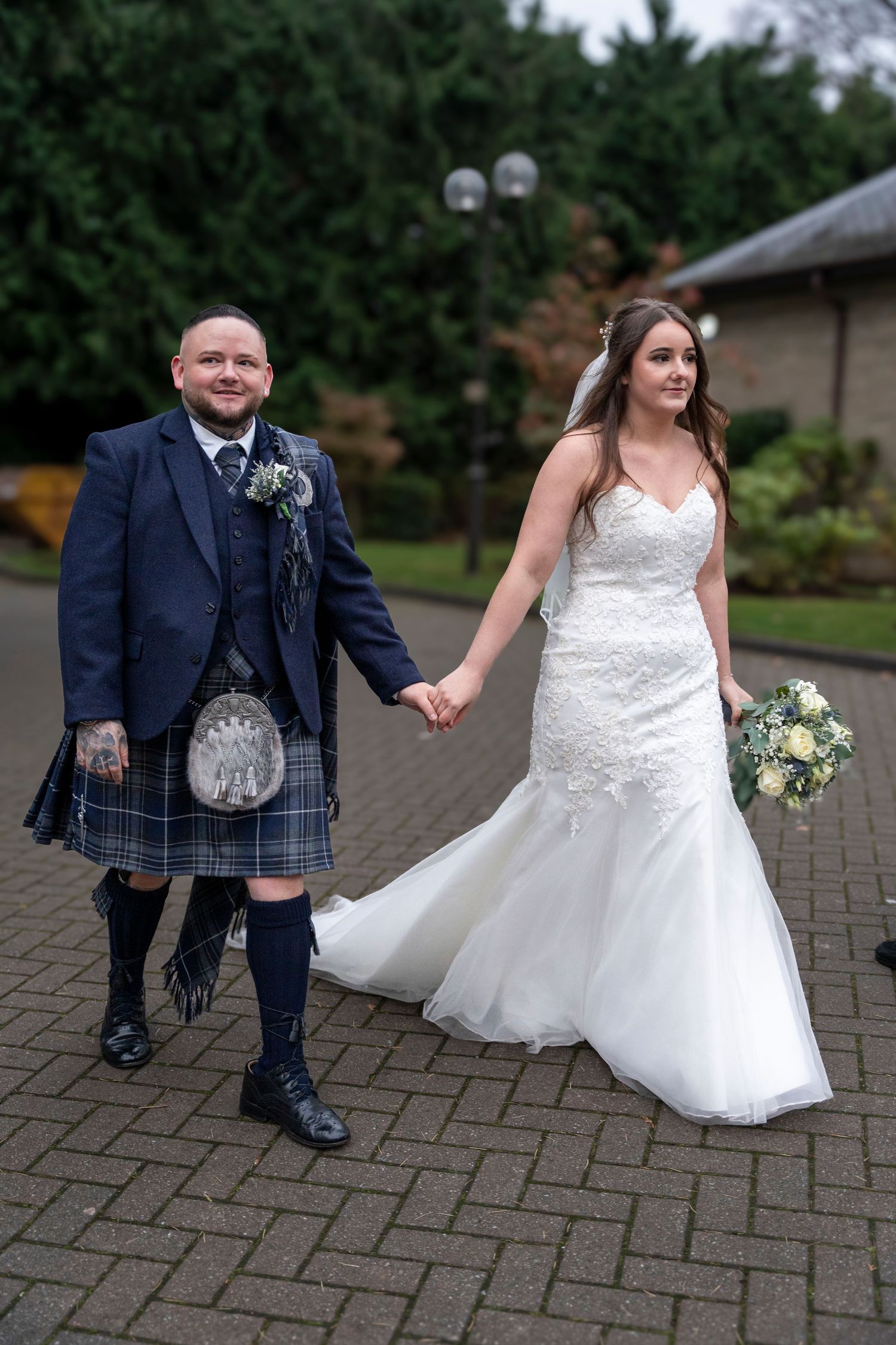 Scottish groom in blue tartan kilt walking hand in hand with bride outdoors. Dave Martin Photographer Northern Ireland