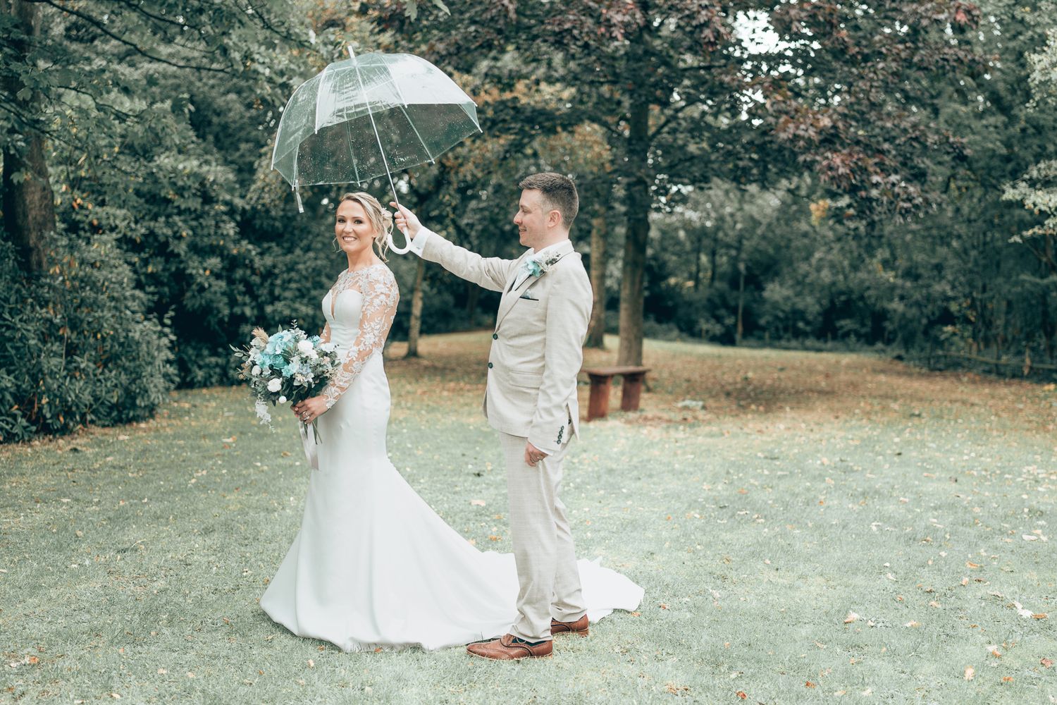 Bride in white gown holds bouquet as groom holds clear umbrella outdoors on wedding day. Dave Martin wedding Photographer NI