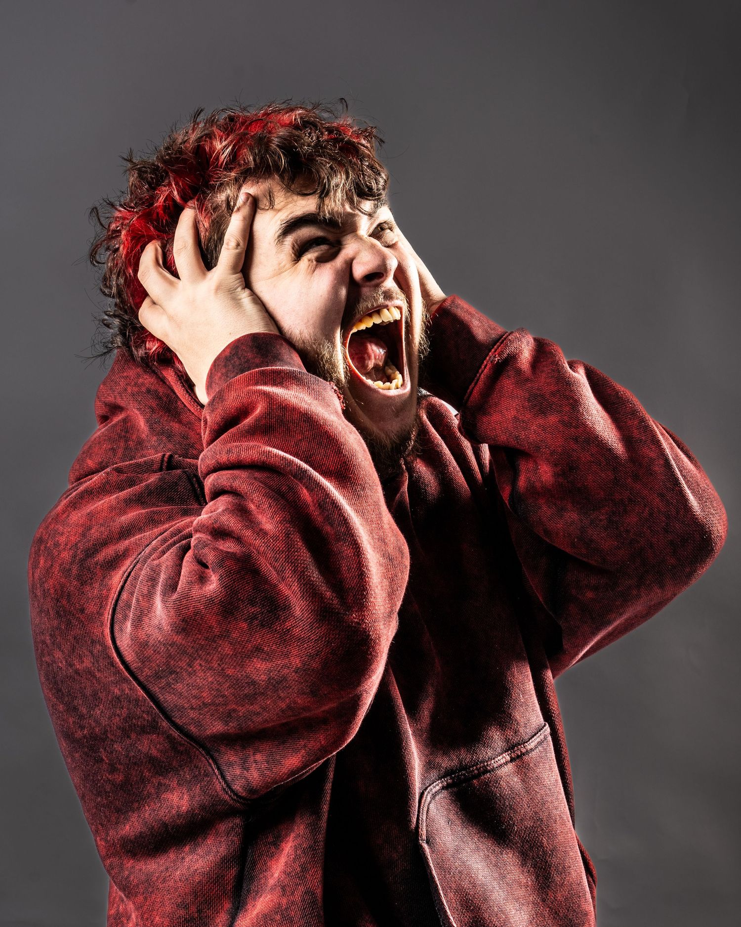 Man in red hoodie screaming with hands on head against gray background, expressing shock or distress. Dave Martin Photography Studio Belfast