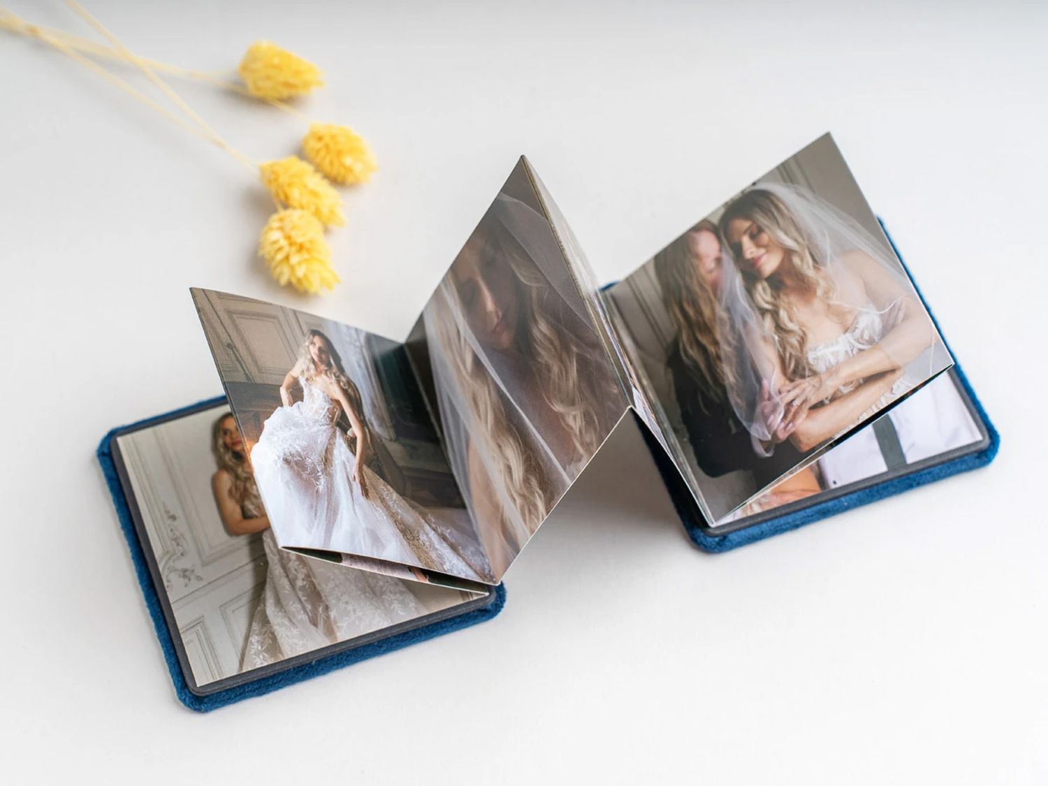 Blue leather accordion photo album open displaying wedding photos, with yellow dried flowers on white background. Northern ireland weddings