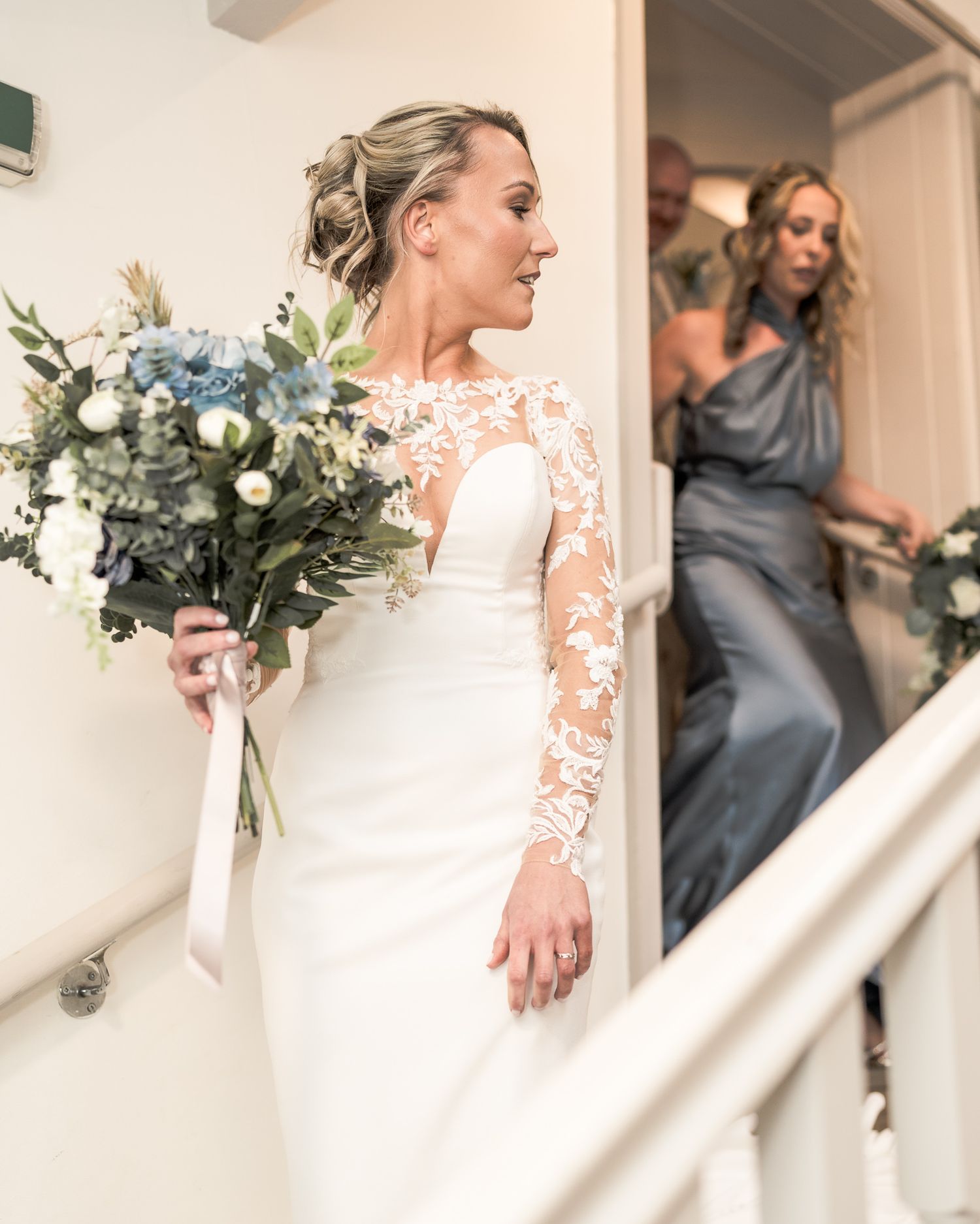 Bride in lace-sleeved white gown holds blue and white bouquet on staircase with bridesmaid in blue dress behind her.