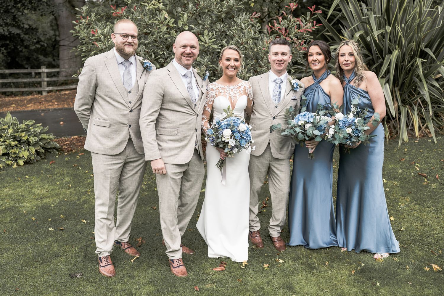 Wedding party posing outdoors, bride in white gown with groomsmen in tan suits and bridesmaids in blue dresses holding floral bouquets.