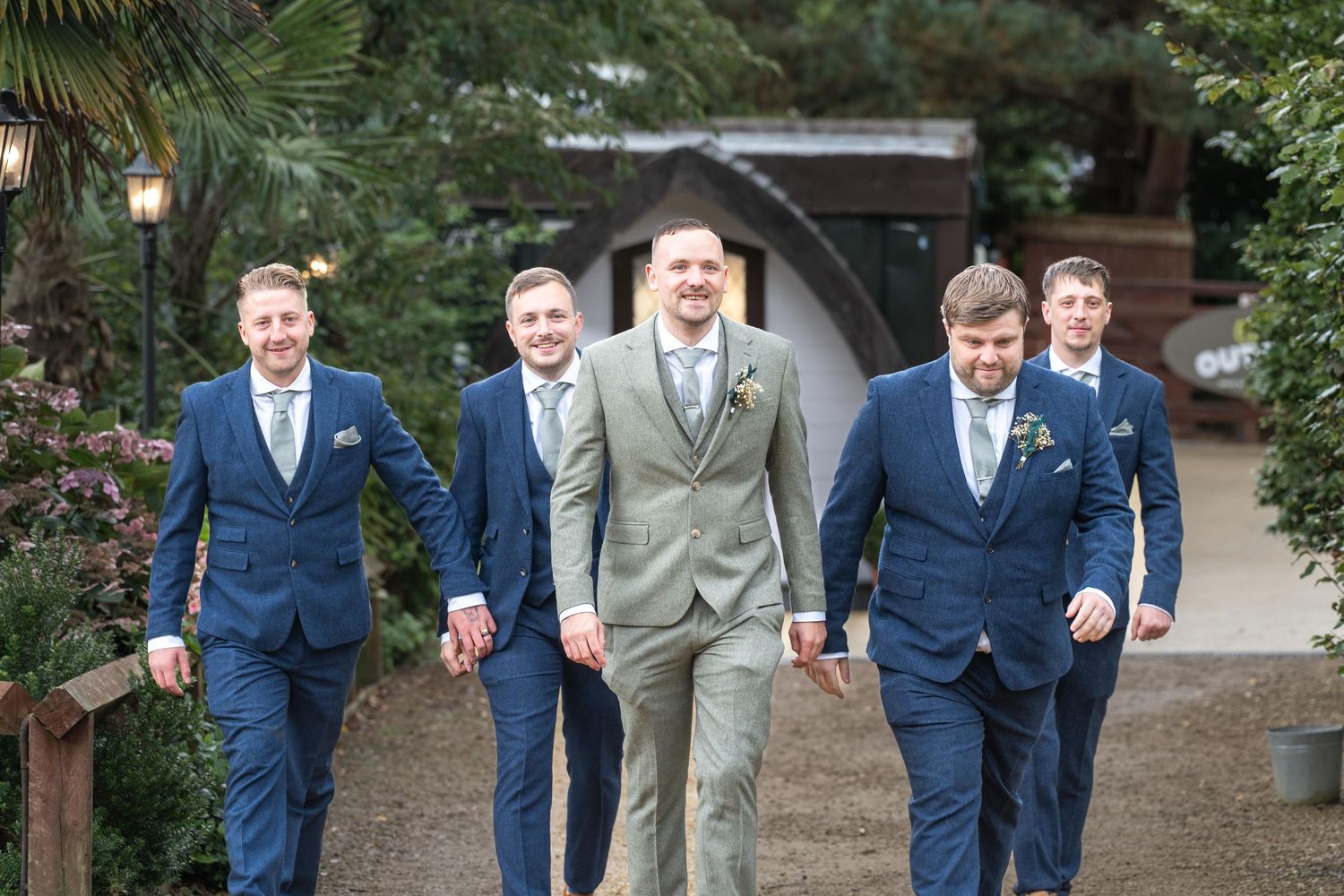 Groom in light gray suit walks with four groomsmen in navy blue suits at an outdoor wedding venue.