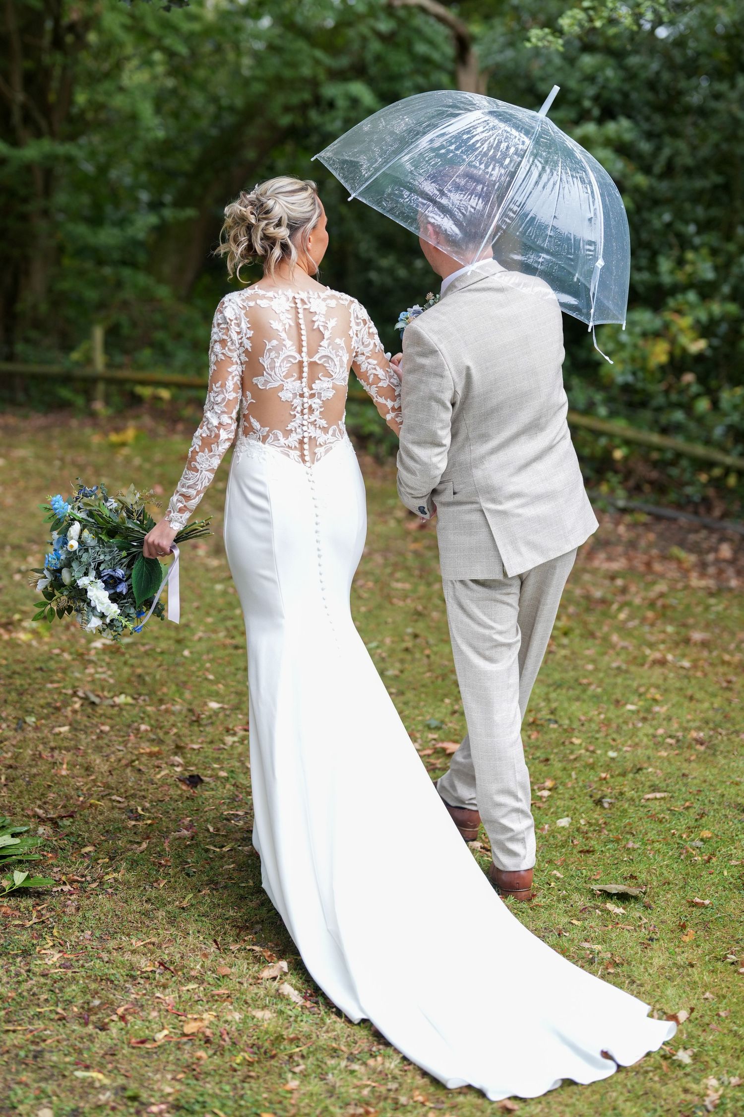 Bride in lace-back gown holding bouquet walks with groom under clear umbrella on green lawn. Northern Ireland Wedding Photographer