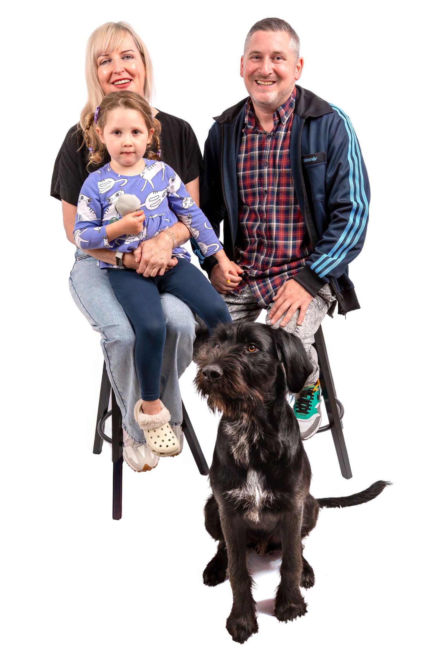 Family portrait with blonde woman, man in plaid shirt, young girl in blue dress, and black dog on white background. Dave Martin Photography
