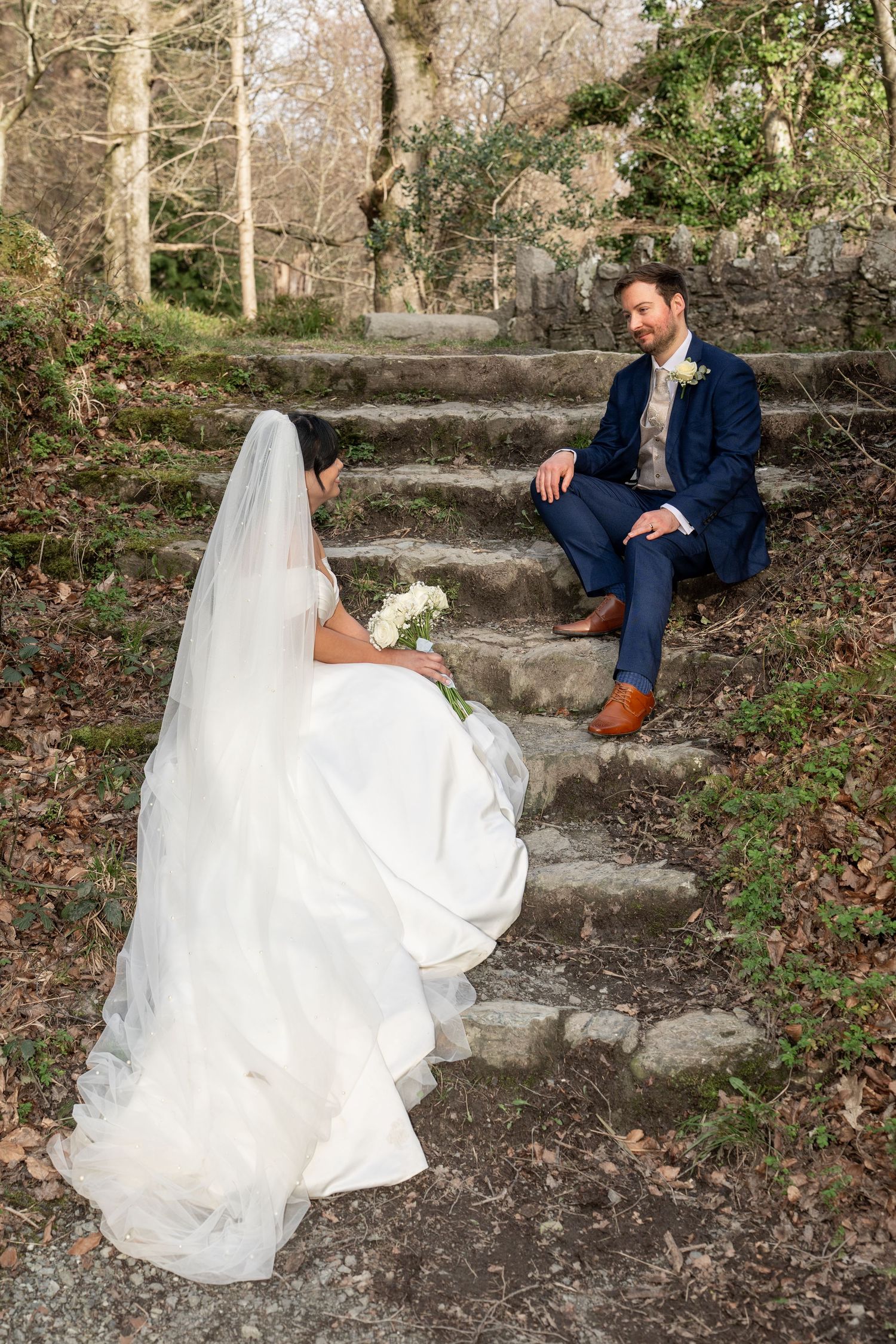 Bride in white gown and veil sitting on stone steps as groom smiles down at her. Dave Martin Photographer Northern Ireland