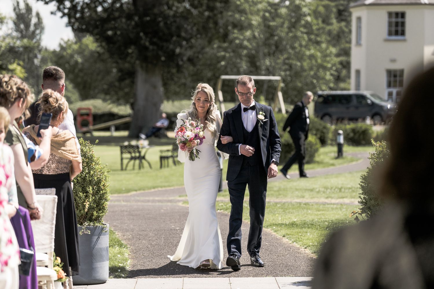Bride and groom walking down outdoor aisle at elegant wedding venue surrounded by guests on a sunny day. Dave Martin wedding Photographer NI