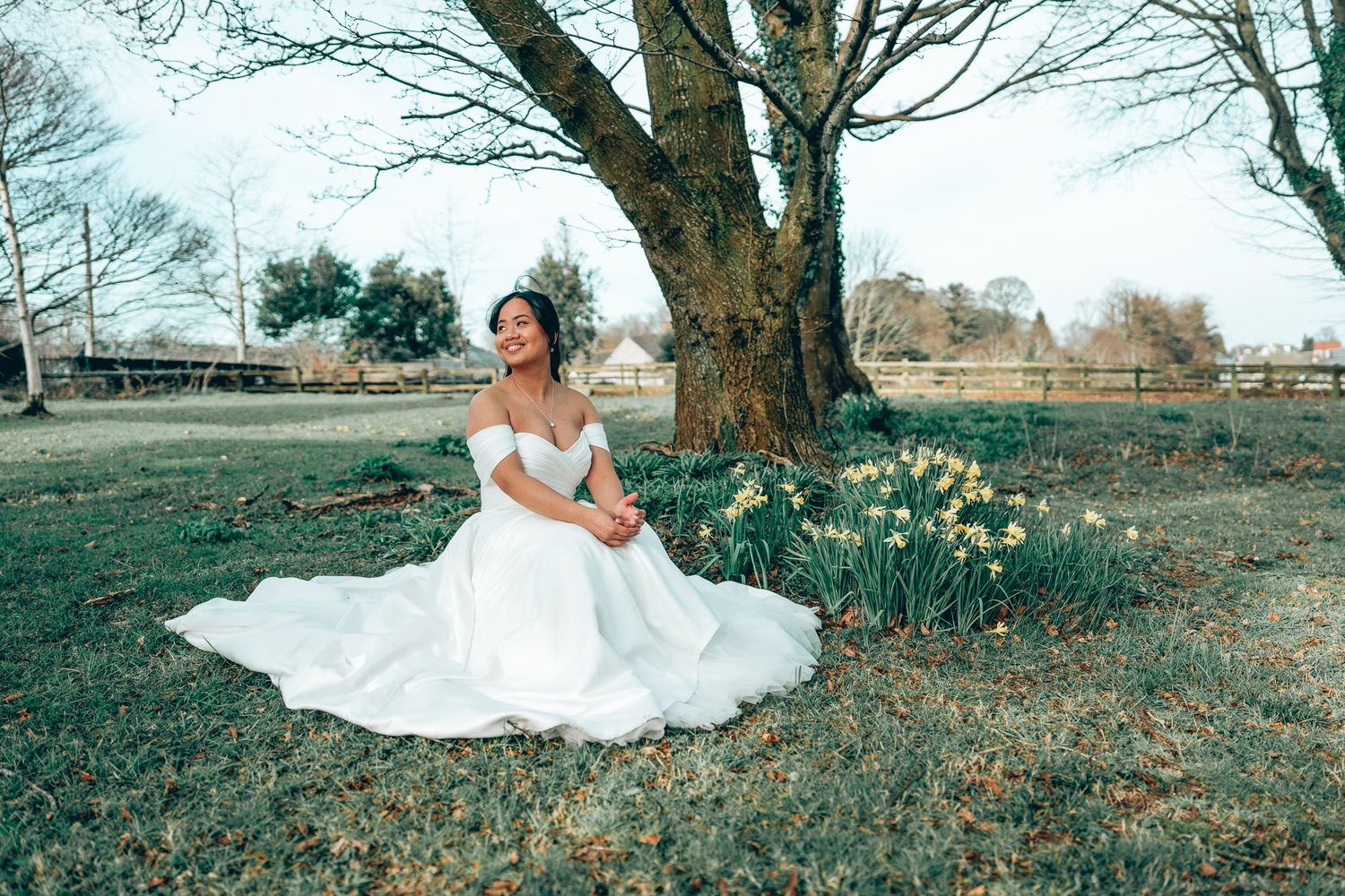 Bride in white off-shoulder gown sitting under a tree with daffodils in a lush green garden. Dave Martin wedding Photographer NI