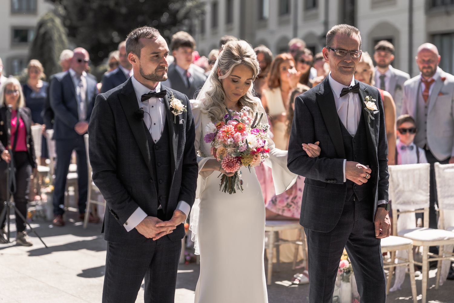 Bride walks down outdoor aisle with two men in tuxedos, holding pink bouquet, guests seated in background. Dave Martin wedding Photographer