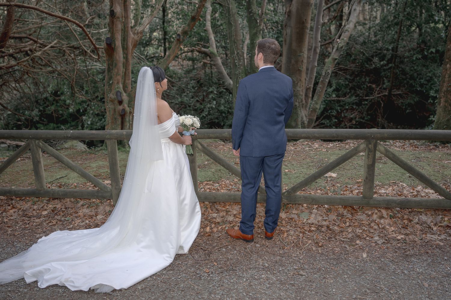 Bride and groom standing together facing a wooden fence surrounded by trees on their wedding day. Dave Martin wedding Photographer NI