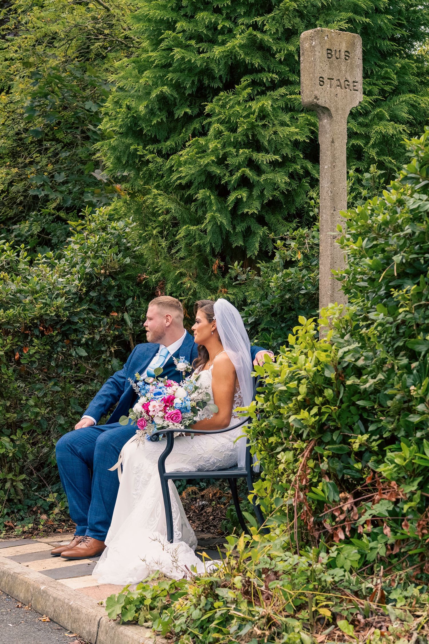 Bride and groom sitting at a vintage bus stop sign surrounded by lush greenery, bride holding a colorful bouquet. By Dave Martin Photography