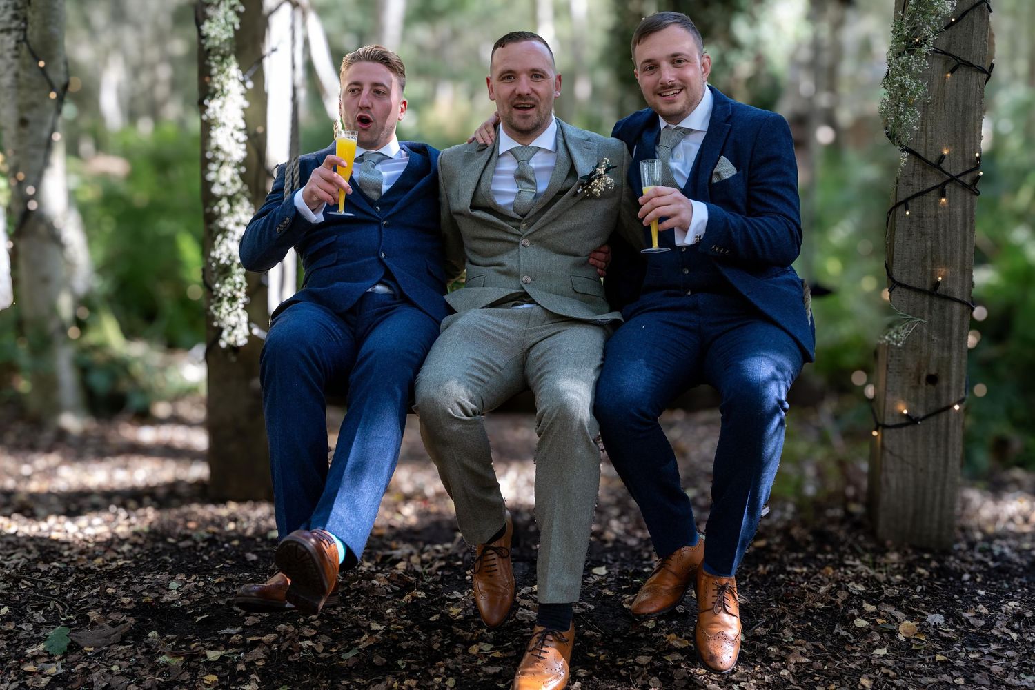 Groom and two groomsmen in navy suits celebrate with champagne on a woodland swing.