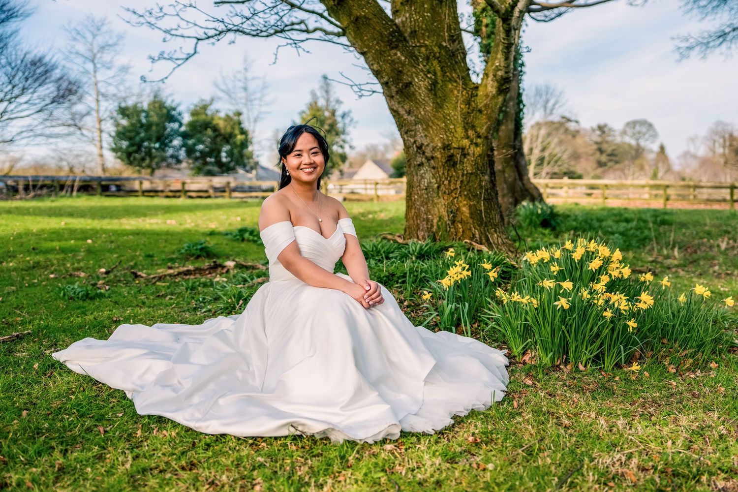 Bride in off-shoulder white gown sits gracefully under tree with daffodils in lush green garden setting.