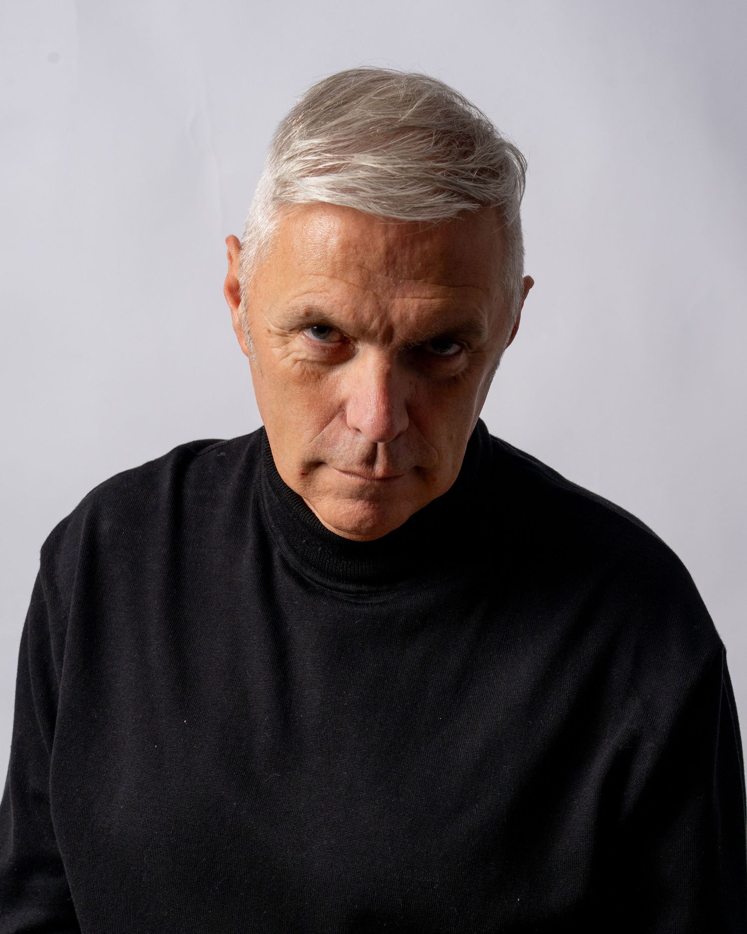 Older man with silver hair wearing a black turtleneck sweater, posing against a light gray background. Dave Martin Photography Studio