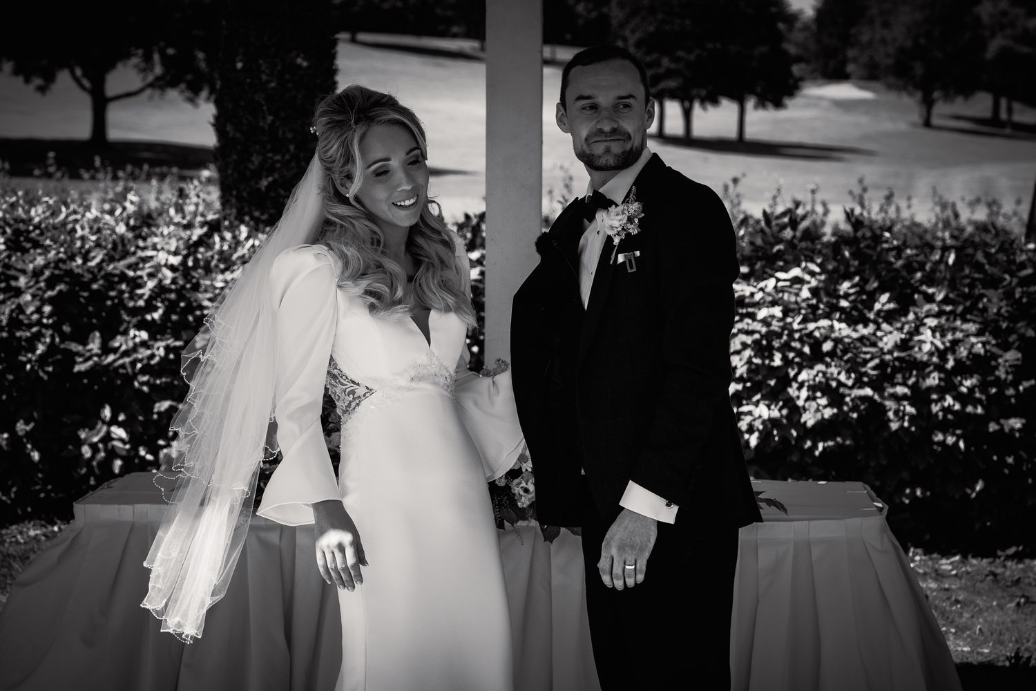 Black and white photo of a smiling bride in a white gown and groom in a dark suit posing together outdoors. Dave Martin wedding Photographer