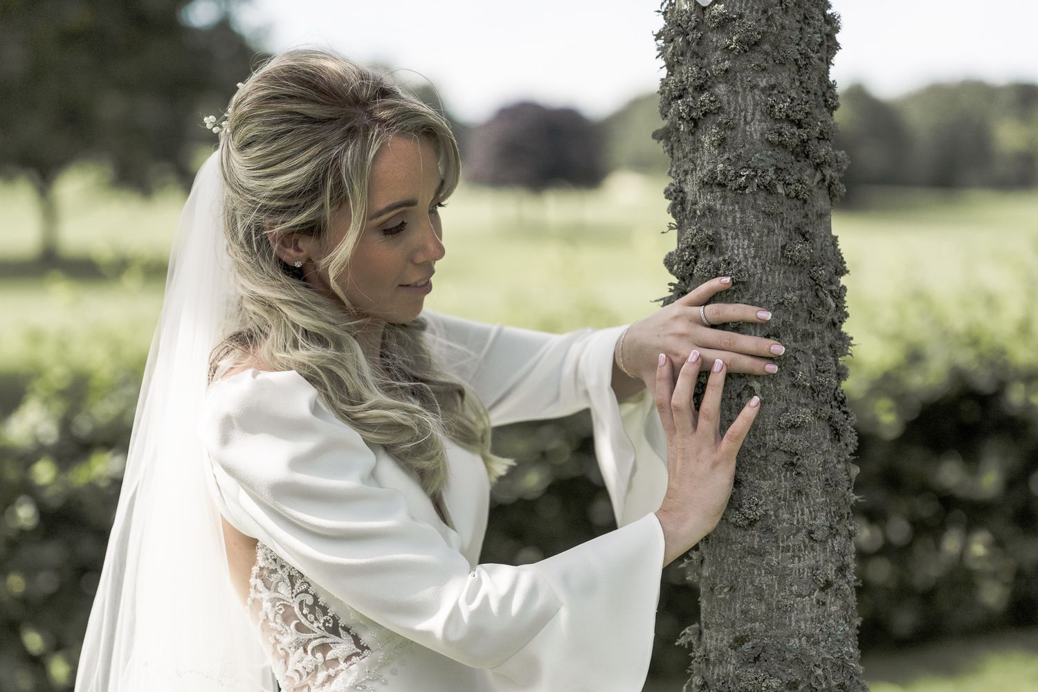 Elegant bride in white lace wedding dress with bell sleeves touches a tree trunk in a garden. Northern ireland weddings