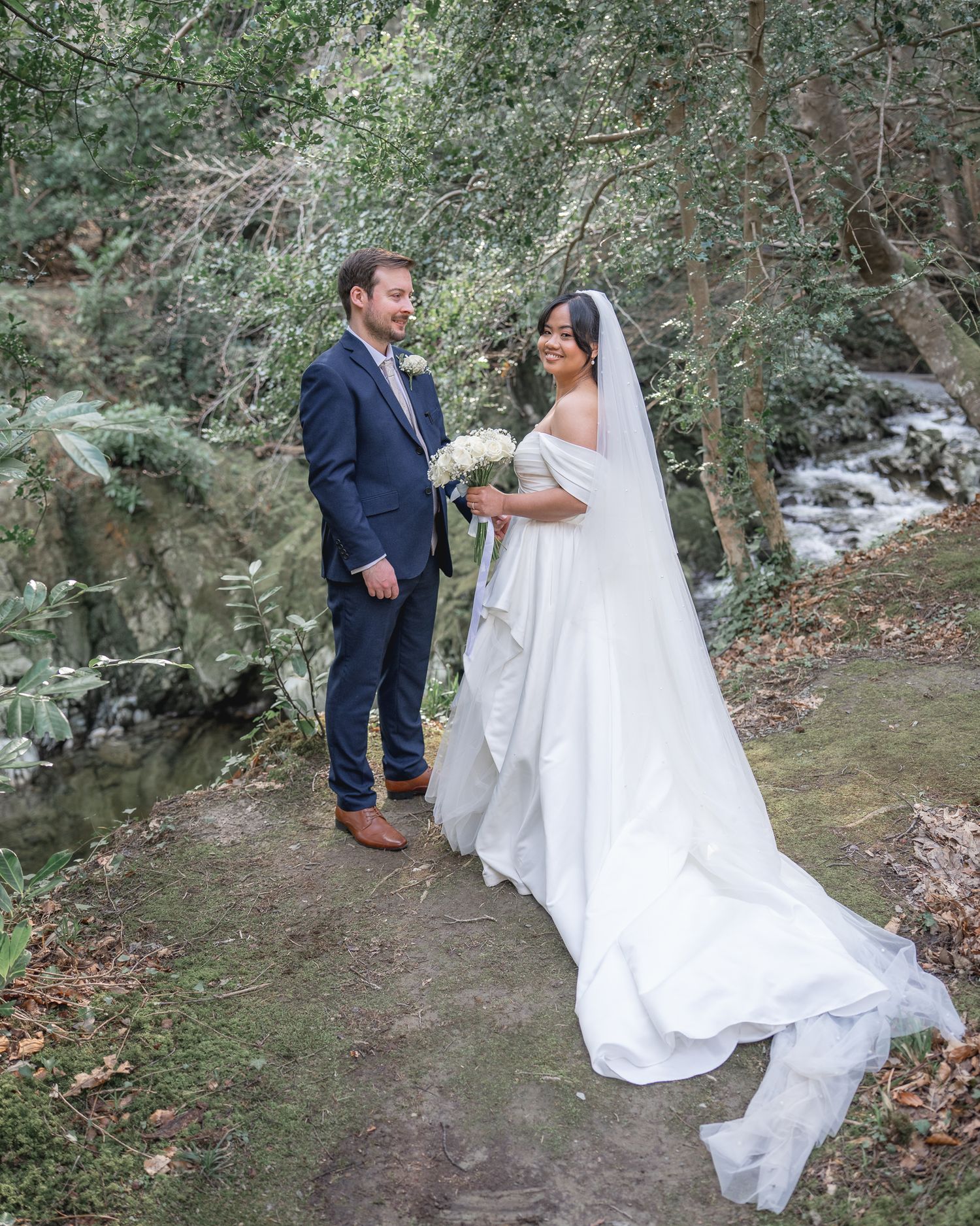 A bride in a white gown with veil and groom in navy suit pose on a forest path near a stream. Dave Martin wedding Photographer NI