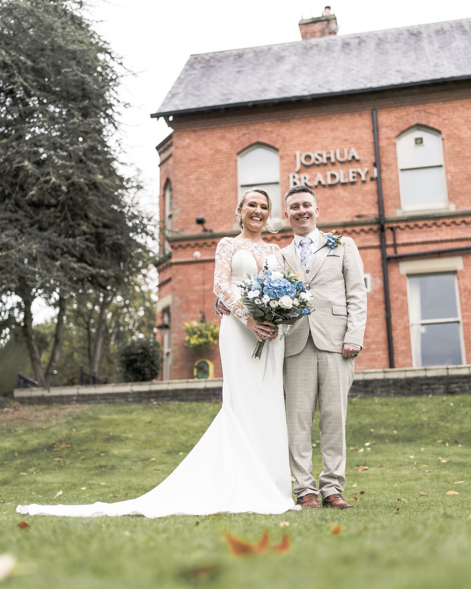 Bride and groom smiling outside Joshua Bradley venue, bride holding blue bouquet in white gown, groom in tan suit.